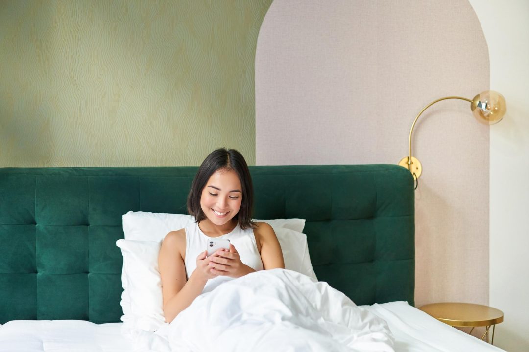 portrait-smiling-asian-woman-lying-bed-looking-smartphone-using-mobile-phone-with-happy-fac